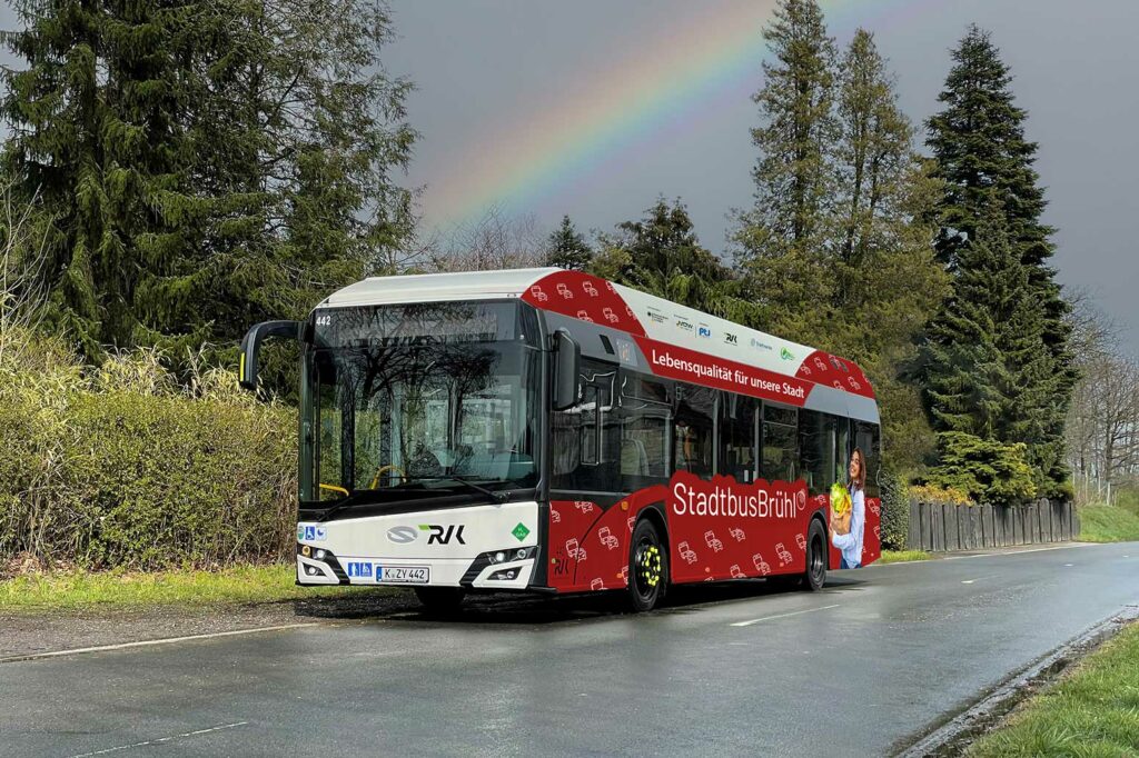 Rot beklebter Stadtwerke Bus auf nasser Straße mit Regenbogen im Hintergrund