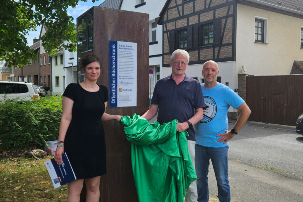3 Personen stehen mit einem grünen Tuch vor einem Bücherschrank