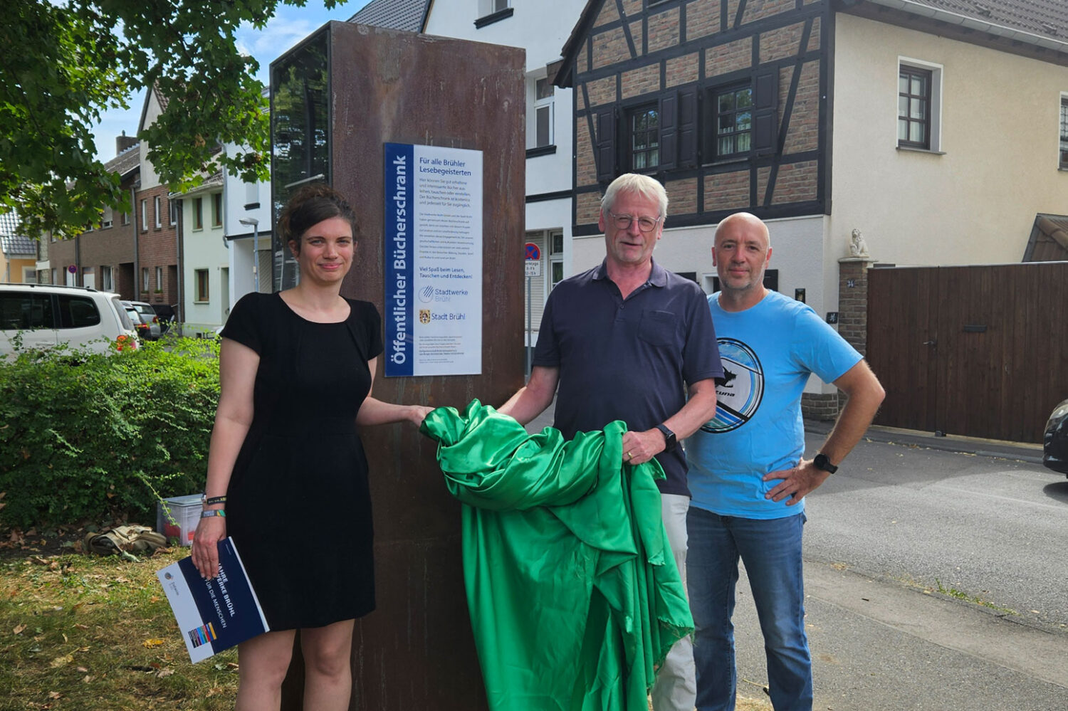 3 Personen stehen mit einem grünen Tuch vor einem Bücherschrank
