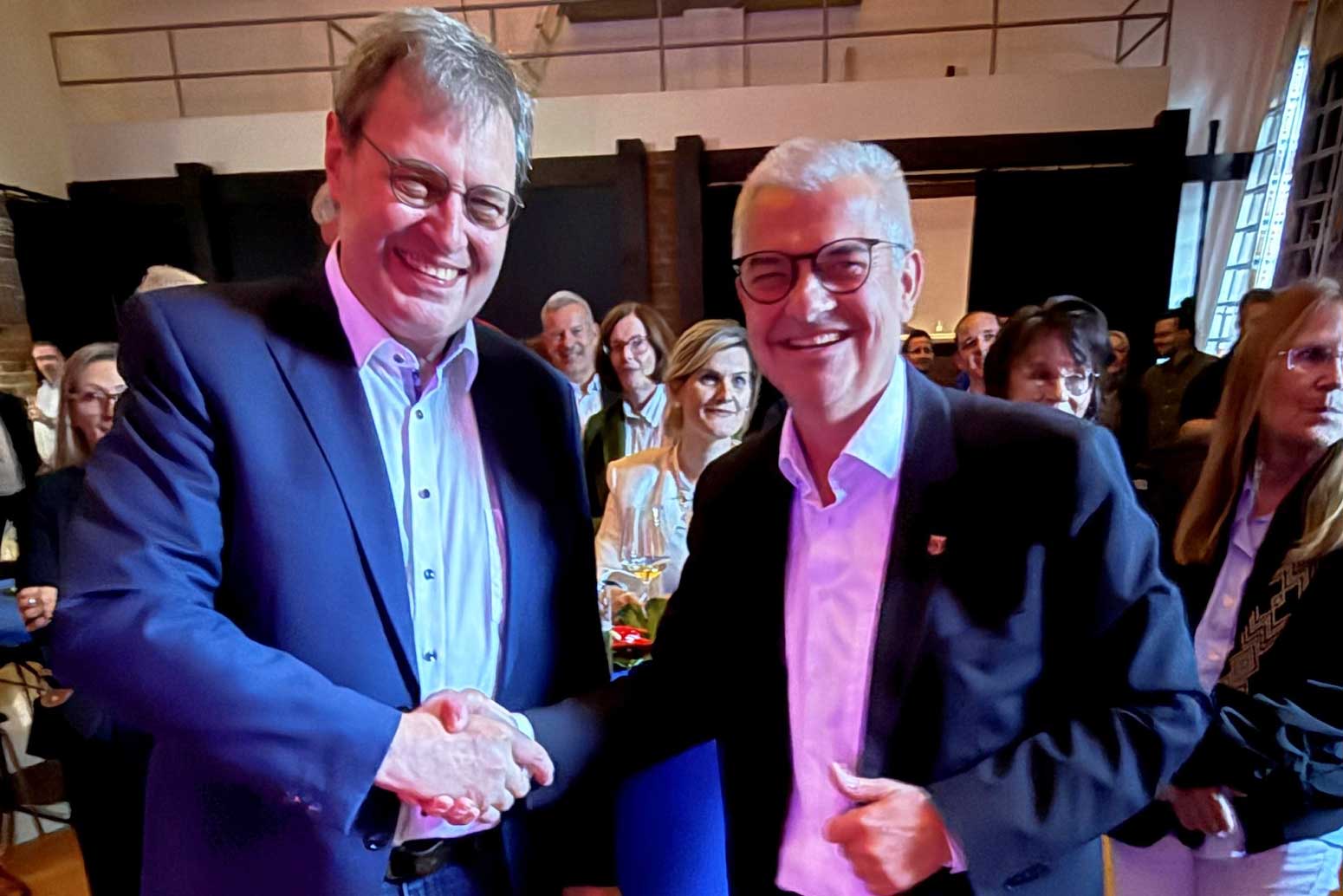 zwei Männer im Festsaal schütteln sich mit Blick in die Kamera die Hände