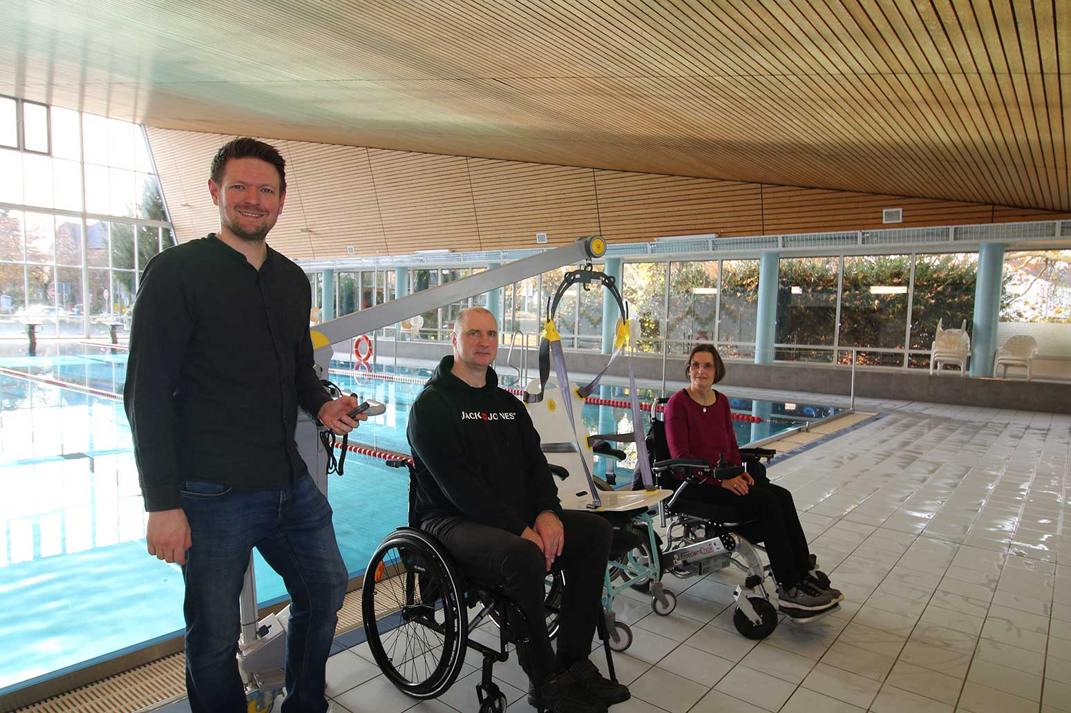 eine gehende Person und zwei Personen im Rollstuhl mit dem neuen Schwimmbadlifter am Sportbecken.